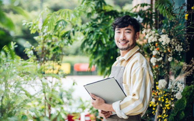 Portrait of confident young Asian male florist, owner of small business flower shop, standing in front of flower shop with clipboard on hand. He is smiling and looking at camera.